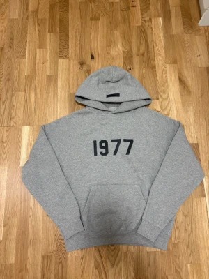 Grå Essentials 1977 hoodie - Snygg grå hoodie från Essentials med svart tryck '1977' på bröstet. Hoodien har en stor huva, magficka och ribbade muddar. Perfekt för en avslappnad streetwear-look. Mjuk bomullsblandning för extra komfort.