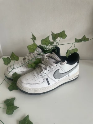 Nike Air Force 1 vita sneakers - Klassiska Nike Air Force 1 sneakers i vitt läder med grå swoosh och svart detalj vid hälen. Skorna har perforerad tå, vita skosnören och svart yttersula. Perfekt för dig som gillar streetwear och tidlös design.