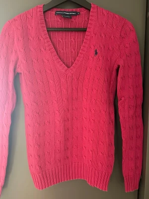 Rosa kabelstickad tröja Ralph Lauren - Snygg rosa kabelstickad tröja från Ralph Lauren Sport med V-ringning och mörkblå logga broderad på bröstet. Tröjan har långa ärmar och ribbade muddar vid ärmslut och nederkant. Tillverkad i mjuk bomull för skön känsla.