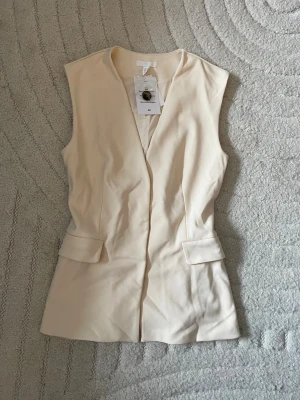 Krämvit väst med fickdetaljer - Stilren krämvit väst i figurnära modell med v-ringning och dekorativa ficklock framtill. Västen har två knappar bak för justering och är tillverkad i ett mjukt tyg som ger en clean look. Perfekt att styla över en skjorta eller t-shirt.