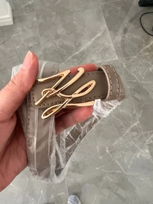 Karl Lagerfeld bälte - Snyggt beige bälte med ett stort, elegant guldspänne i form av ett stiliserat bokstavsmotiv. Bältet har en clean look och passar perfekt till jeans eller byxor för att lyfta din outfit.
