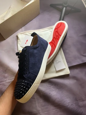 Blåa Louboutin sneakers med nitar - Snygga mörkblå sneakers från Christian Louboutin i mocka med svarta nitar på tån och klassisk röd sula. Skorna har vita sulor och svarta detaljer, perfekt för dig som vill sticka ut med en lyxig och edgy stil.