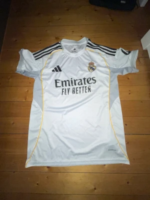 Real Madrid matchtröja Adidas vit - Snygg vit Real Madrid matchtröja från Adidas med svarta detaljer och gula linjer. Tröjan har klubbmärke, Adidas-logga och Emirates Fly Better-tryck på bröstet. Tillverkad i lätt och ventilerande material, perfekt för fotboll eller träning.Pris går att diskuteras
