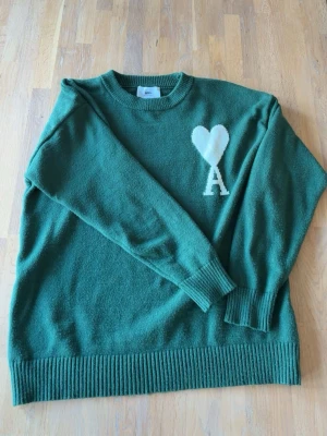 Grön Ami Paris Tröja - Mörkgrön stickad tröja från Ami Paris med rund halsringning och ribbade muddar. På bröstet finns ett vitt hjärta och bokstaven A. Tröjan har en avslappnad passform och är perfekt för lager-på-lager. Materialet är otroligt skönt