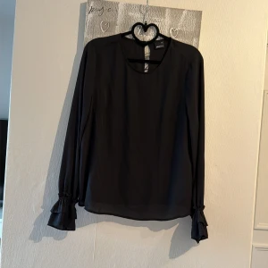Svart blus med volangärm från Gina Tricot - Stilren svart blus från Gina Tricot i ett tunt, lätt material. Blusen har långa ärmar med volangdetalj vid ärmslut och en rund halsringning. Perfekt för en trendig och elegant look.