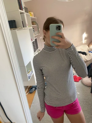 Randig polotopp i svart och vitt - Säljer en långärmad polotopp med smala svarta och vita ränder. Toppen har en hög krage och är i en slim passform som sitter snyggt mot kroppen. Perfekt att styla med jeans eller kjol för en clean look.