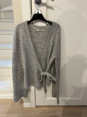 Grå omlottkofta från Gina Tricot - Mysig grå omlottkofta från Gina Tricot i mjukt stickat material. Koftan har långa ärmar, ribbade kanter och knyts i midjan med ett band. Perfekt för lager på lager och enkel att styla till olika outfits.