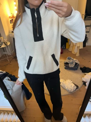 Vit teddytröja med svarta detaljer - Teddytröja i vitt med svarta detaljer vid dragkedja och fickor. Hög krage och halv dragkedja framtill. Tillverkad i mjukt, fluffigt material som ger en avslappnad och trendig look. Långa ärmar och två fickor framtill.