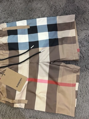 Rutig Burberry shorts med snörning - Snygga shorts från Burberry med klassiskt rutigt mönster i beige, svart, vit, blå och röd. Shortsen har snörning i midjan och är perfekta för sommaren. Tillverkade i ett lätt och bekvämt material som passar till alla tillfällen.