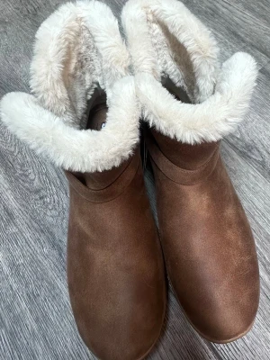 Bruna boots   - Bruna boots i mockaimitation med vitt fluffigt fuskpälsfoder. Stövlarna har rund tå, dekorativ rem på sidan och en rejäl vit sula som ger bra grepp. Perfekta för kalla dagar när du vill ha både stil och värme. De är lätta att ta av och på. Dem är i nyskick, endast testade. 