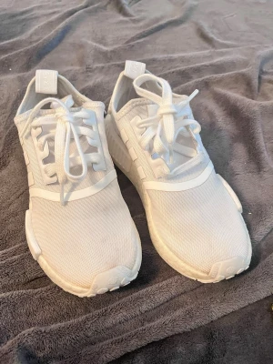 Adidias NMD - Säljer ett par helvita Adidas NMD sneakers med klassiska tre ränder på sidan. Skorna har ovandel i mesh och syntet, samt en bekväm Boost-sula. Lågt skaft och snörning framtill. Perfekta för dig som gillar stilrena och sportiga sneakers.
