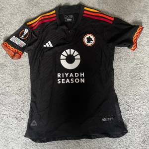 AS Roma matchtröja Dybala 21 - Snygg AS Roma matchtröja med Paulo Dybala på ryggen. En väldigt detaljrik tröja som också har Europa League patch på ena tröjärmen. Den är i ”player version”, som är den sorts material spelarna har under match. Tröjan är i storlek M, är knappt använd och är alltså i perfekt skick.