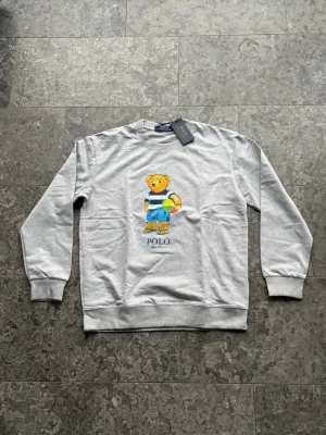 Grå sweatshirt med Polo Bear från Ralph Lauren - Grå sweatshirt från Polo Ralph Lauren med det ikoniska Polo Bear-motivet på bröstet. Björnen har på sig blå shorts, randig tröja och håller en färgglad badboll. Tröjan har rund hals och ribbade muddar. Perfekt för en avslappnad och trendig look.