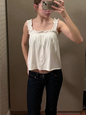 Vit volanglinnetopp med breda axelband - Söt vit linnetopp med breda axelband och volangdetaljer. Toppen har en lös passform och smockad kant upptill för extra komfort. Perfekt för varma dagar och enkel att styla med jeans eller kjol.