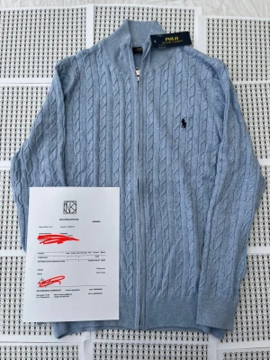 Ljusblå kabelstickad kofta Polo Ralph Lauren - Kabelstickad ljusblå kofta från Polo Ralph Lauren med dragkedja framtill och ribbade muddar. Klassisk broderad logga på bröstet och hög krage. Perfekt för dig som gillar stilrena och tidlösa plagg.