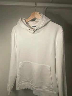 Vit hoodie från J.Lindeberg - Säljer en stilren vit hoodie från J.Lindeberg med broderad logga på bröstet. Hoodien har en klassisk känguruficka framtill och justerbar huva med snörning. Tillverkad i mjukt bomullsmaterial som är skönt att bära.