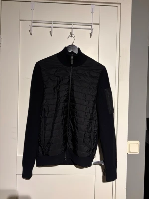 Svart cardiganjacka j-Lindeberg  - Stilren svart cardiganjacka med quiltad front och stickade ärmar. Jackan har hög krage, hel dragkedja och en ficka med dragkedja på ena ärmen. Materialmix av vadderad polyester framtill och stickad bomull eller ull på ärmar och rygg. På bild 5 ser man en lagning i armen. Den syns inte när man använder den eftersom den är på insidan och bra sydd.