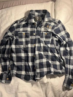 Blårutig skjorta från H&M L.O.G.G. - Snygg blårutig skjorta från H&M:s L.O.G.G.-kollektion med två bröstfickor och broderad detalj på ena sidan. Skjortan har klassisk krage, knappar i kontrastfärg och långa ärmar. Perfekt för en avslappnad och trendig stil.