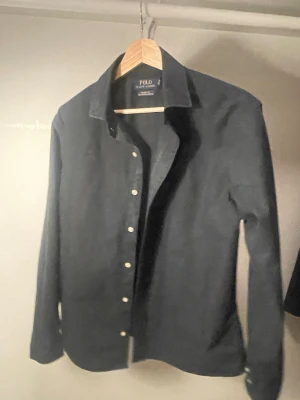 Svart skjorta från Polo Ralph Lauren - Stilren svart skjorta från Polo Ralph Lauren med klassisk krage och vita knappar. Skjortan är i bomull och har en tidlös design som passar perfekt till många olika looks. Classic fit-modell som ger en avslappnad men snygg siluett.