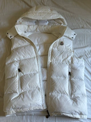 Vit dunväst från Moncler med huva - Snygg vit dunväst från Moncler med avtagbar huva och två dragkedjeförsedda fickor. Västen har klassisk Moncler-logga på bröstet och coolt serietryck på insidan. Tillverkad i glansigt material och fylld med dun för extra värme. Perfekt för lager på lager under kalla dagar. lite skitig i luvan, skriv för mer info kvitto,  bilder ,osv