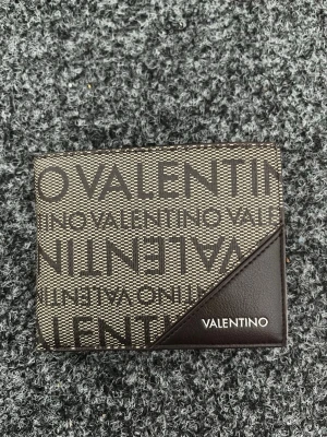 Valentino plånbok - Snygg plånbok från Valentino med grafiskt logomönster i svart och beige. Plånboken har en unik design med en diagonal läderdetalj i hörnet där Valentino-loggan är tryckt. Tillverkad i tåligt material och passar perfekt för kort och sedlar. Köpt för 499kr!