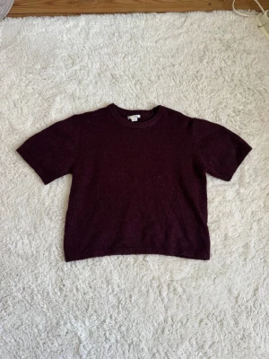 Vinröd stickad t-shirt - Kortärmad stickad t-shirt i vinrött med rund halsringning. Tröjan har en rak passform och är gjord i ett mjukt, mysigt material som passar perfekt till lager på lager. Enkel och stilren design utan synliga detaljer eller mönster. I storlek 158/164 men passar XS