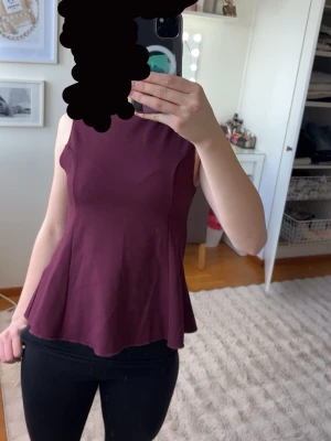 Ärmlös vinröd peplumtop från H&M - Säljer en snygg ärmlös peplumtop från H&M i vinrött. Toppen har rund halsringning och markerad midja som ger en fin siluett. Tillverkad i ett mjukt, stretchigt material som sitter bekvämt. Perfekt att styla med jeans eller kjol.