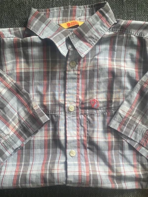 Rutig skjorta från Fjällräven - Snygg rutig skjorta från Fjällräven i grå, röd och vit färgskala. Skjortan har klassisk krage, knappar framtill och ett broderat rött Fjällräven-märke på bröstet. Tillverkad i mjuk bomull, perfekt för en avslappnad stil.