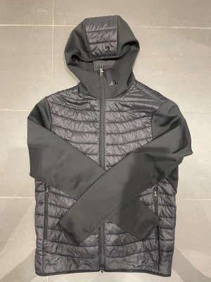 J.lindeberg cardigan - Snygg svart vindjacka med quiltad front och rygg samt släta ärmar. Jackan har hel dragkedja, två sidofickor med dragkedja och en fast huva. Materialet är en mix av lättviktsvaddering och softshell-tyg för extra komfort och stil.