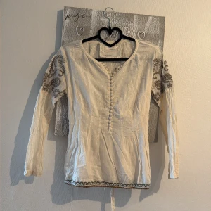 Vit boho-blus med broderi - Säljer en vit boho-inspirerad blus med långa ärmar och dekorativa knappar framtill från Odd Molly. Blusen har fina bruna broderier på ärmarna och ett band i midjan som knyts bak. Materialet är tunt och lätt, perfekt för en avslappnad stil.