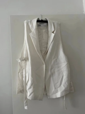 Vit väst med knyt från ZARA - Stilren vit väst från ZARA med klassisk krage och knytband i sidan. Västen har en knapp framtill och är tillverkad i ett mjukt tyg som ger en clean look. Perfekt att styla över en skjorta eller t-shirt för en trendig vibe. Använd en gång till en fest.