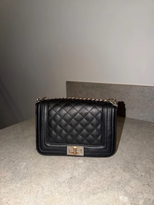 Svart handväska med kedja - Svart handväska med silverfärgad kedjerem och metallspänne framtill.