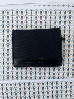 Svart plånbok Tiger of Sweden - Snygg svart plånbok från Tiger of Sweden i textil och läderdetaljer. Plånboken har flera kortfack och ett genomskinligt fack för ID eller körkort. Klassisk och stilren design med logga på framsidan.