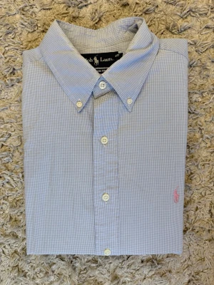 Blårutig skjorta från Ralph Lauren - Klassisk blårutig skjorta från Ralph Lauren med lång ärm och knappar framtill. Skjortan har en liten rosa broderad logga på bröstet och är tillverkad i mjuk bomull. Perfekt för en clean och stilren look.