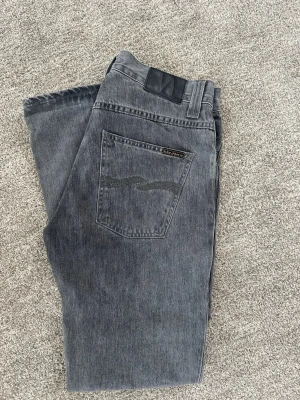 Grå Nudie Jeans Grim Tim - Säljer ett par grå jeans från Nudie Jeans med klassisk femficksmodell och raka ben. Jeansen har en snygg tvättad look och detaljerad söm på bakfickorna. Tillverkade i slitstark denim, slim fit modell Grim Tim. Storlek 31 i midja, 32 i längd.