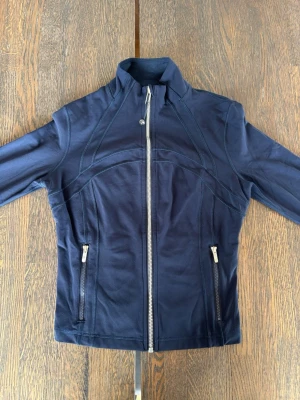 NY Marinblå jacka från Lululemon storlek S/6 - Snygg marinblå jacka från Lululemon med figurnära passform och silverfärgade dragkedjor. Jackan har hög krage, långa ärmar och två fickor med dragkedja. Perfekt för dig som gillar sportig stil och vill ha något bekvämt och stilrent. Har haft på mig den bara en gång så den är helt ny. Pris i butik är 1400 SEK.