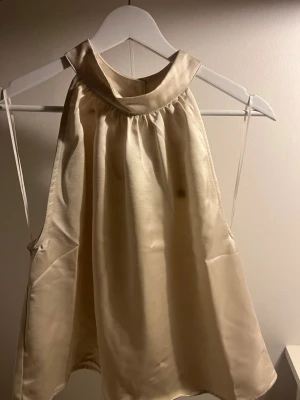 Champagnefärgad halternecktop Bik Bok - Snygg champagnefärgad halternecktop från Bik Bok med glansigt satintyg och bred krage. Toppen har en lös passform och knyts i nacken. Perfekt för att skapa en chic och trendig look.