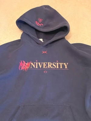 ABN hoodie  - Snygg mörkblå hoodie  med trycket 'UNIVERSITY' i gult och rött på bröstet samt coola grafiska detaljer. Huva med broderad logga och stor magficka framtill. Perfekt för dig som gillar streetwear och vill sticka ut lite extra.