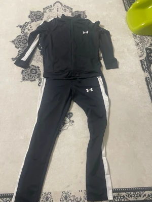 Svart träningsset från Under Armour - Säljer ett svart träningsset från Under Armour med vita detaljer längs ärmar och ben. Setet består av en zip-jacka och matchande byxor, båda med Under Armour-logga på bröst och lår. Materialet är mjukt och stretchigt, perfekt för träning eller sport.
