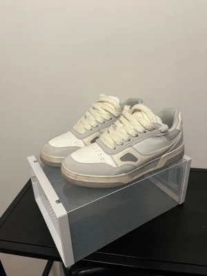 Vita och grå sneakers med chunky sula - Snygga sneakers i vitt och ljusgrått med breda, tjocka snören och chunky sula. Ovandelen är i en mix av läder och mesh för extra komfort och stil. Perfekta för dig som gillar en clean och modern look.