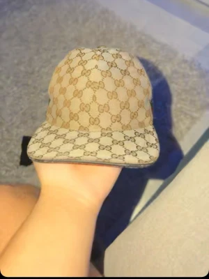 Beige GG-mönstrad keps från Gucci - Säljer en beige keps från Gucci med det klassiska GG-monogrammet över hela kepsen. Kepsen har böjd skärm och justerbart band bak. Materialet är canvas med läderdetaljer. Perfekt för dig som vill ha en lyxig och trendig accessoar.Storlek M säljs billigt 