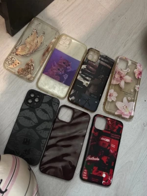 7-pack mobilskal med olika motiv iphone 11 pro max - Säljer ett 7-pack mobilskal till iPhone med olika motiv och färger. Här finns skal med blommor, fjärilar, kattmotiv, mörkröda marmorvibbar, svarta läppar och ett rött collage. Materialet är mestadels plast och silikon. Perfekt för dig som vill variera stilen på mobilen!