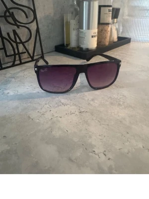 Svarta Ray-Ban solglasögon - Snygga svarta solglasögon från Ray-Ban med rektangulär form och mörka tonade glas. Klassisk design som passar till de flesta outfits. Bågarna är i plast och har Ray-Ban-loggan på linsen.