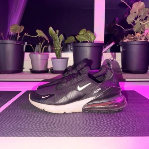 Nike Air Max 270 svarta sneakers - Svarta Nike Air Max 270 sneakers med vit sula och synlig Air-enhet i hälen. Skorna har mesh-ovandel, svarta skosnören och små vita Nike-loggor på sidan och tån. Sportig design med dragflik bak och diskreta detaljer. Perfekta för en streetwear-look.