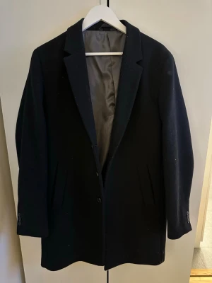 Svart klassisk ullrock | Jack & Jones - Stilren svart rock från Jack & Jones med enkelknäppning och två fickor framtill. Rocken har klassisk krage och slät design, perfekt för en clean och tidlös look. Fodrad insida för extra komfort. Passar dig som gillar minimalistisk och elegant stil.