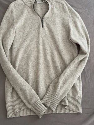 Beige stickad tröja med halv dragkedja - Säljer en beige stickad tröja från Kappahl  med halv dragkedja vid halsen. Tröjan har långa ärmar, ribbstickade muddar och nederkant samt en mjuk, lite melerad yta. Perfekt för dig som gillar stilrena och klassiska plagg.