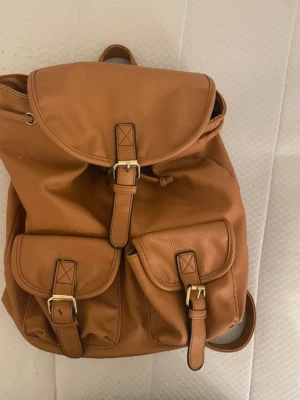 Karamellbrun ryggsäck med spännen - Snygg karamellbrun ryggsäck i fejkläder med två framfickor och guldfärgade spännen. Ryggsäcken har justerbara axelband och dragsko under locket. Perfekt för dig som vill ha en stilren och praktisk väska till skolan eller stan.