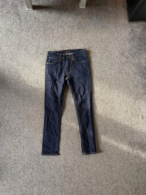 Mörkblåa jeans från Nudie - Säljer ett par mörkblå jeans från Nudie Jeans med klassiska fem fickor och kontrastsömmar. Jeansen har en smal passform och är tillverkade i denim med snygga detaljer på bakfickorna. Perfekta för en stilren och avslappnad look.