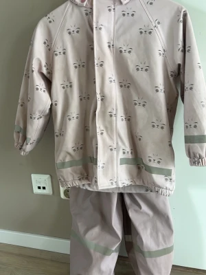 Ljusrosa set från Mywear Young 122-128 - Tvådelat set i ljusrosa med sött kattansikte-mönster. Jackan har dragkedja, huva och reflexdetaljer på ärmar och nederkant. Byxorna har justerbara hängslen, reflexband och mjukt fleecefoder. Perfekt för utomhuslek under kyliga dagar.