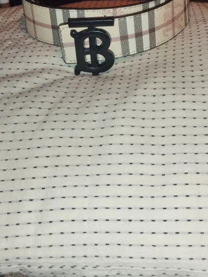 Rutmönstrat Burberry bälte - Snyggt bälte från Burberry med klassiskt rutmönster i beige, svart och brunt. Bältet har ett stort svart metallspänne med Burberrys ikoniska monogram. Tillverkat i skinn och passar perfekt för att lyfta vilken outfit som helst. Den är helt ny. Kan användas i två sidor både beige och svart.